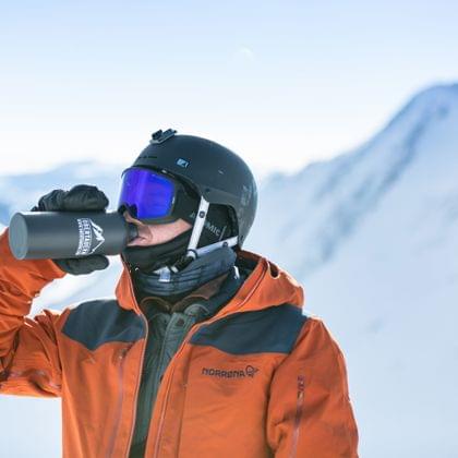 Skifahrer in orangefarbener Jacke trinkt aus Flasche vor verschneiten Alpen